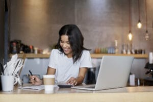 A smiling woman works at her laptop and writes on a notepad.