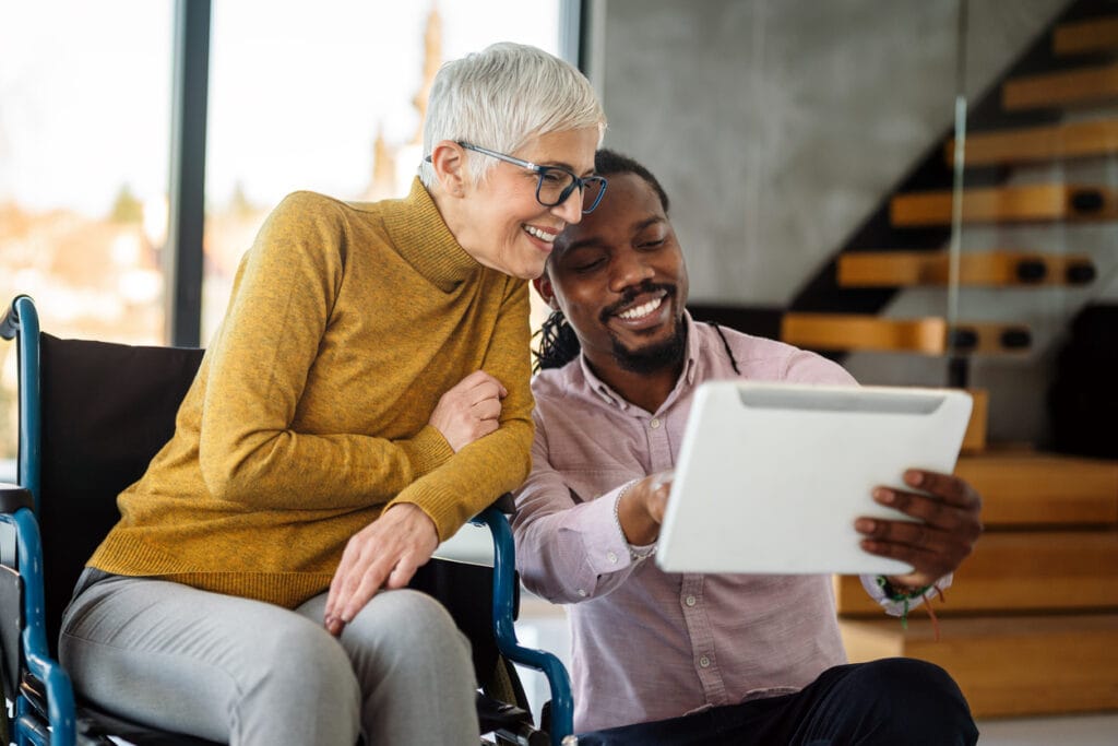 A man holds a tablet while a woman in a wheelchair leans over his shoulder. Website accessibility is a non-negotiable for all websites.