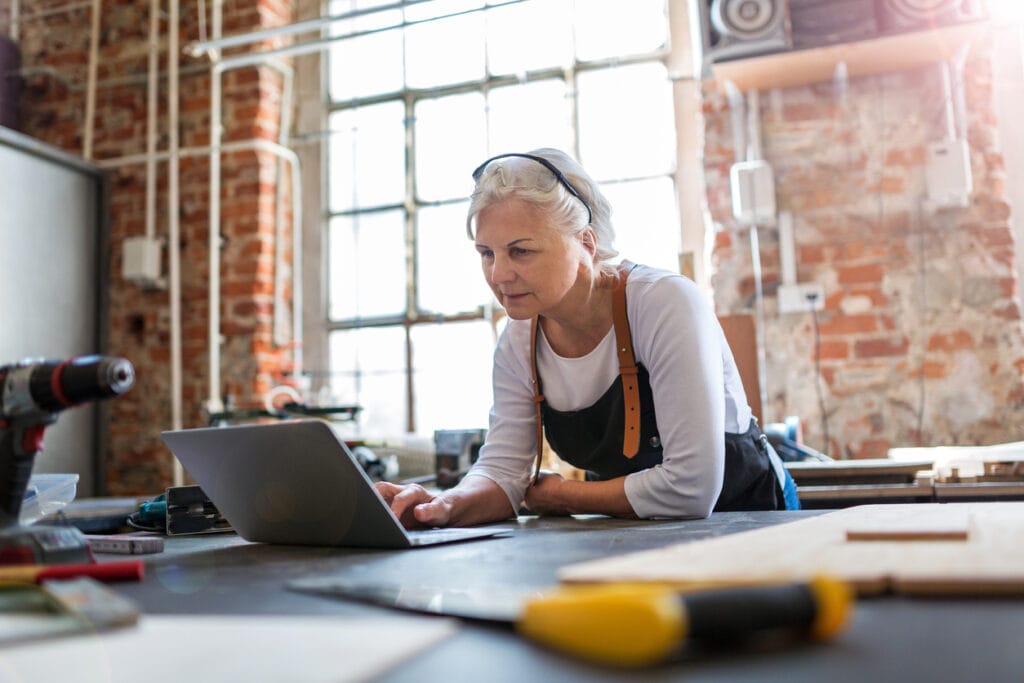 A woman works on her laptop in a brick building, bending over a table.