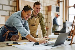 Warm toned portrait of animated carpenters using laptop and discussing furniture designs in workshop The Valley List offers small business website design services.