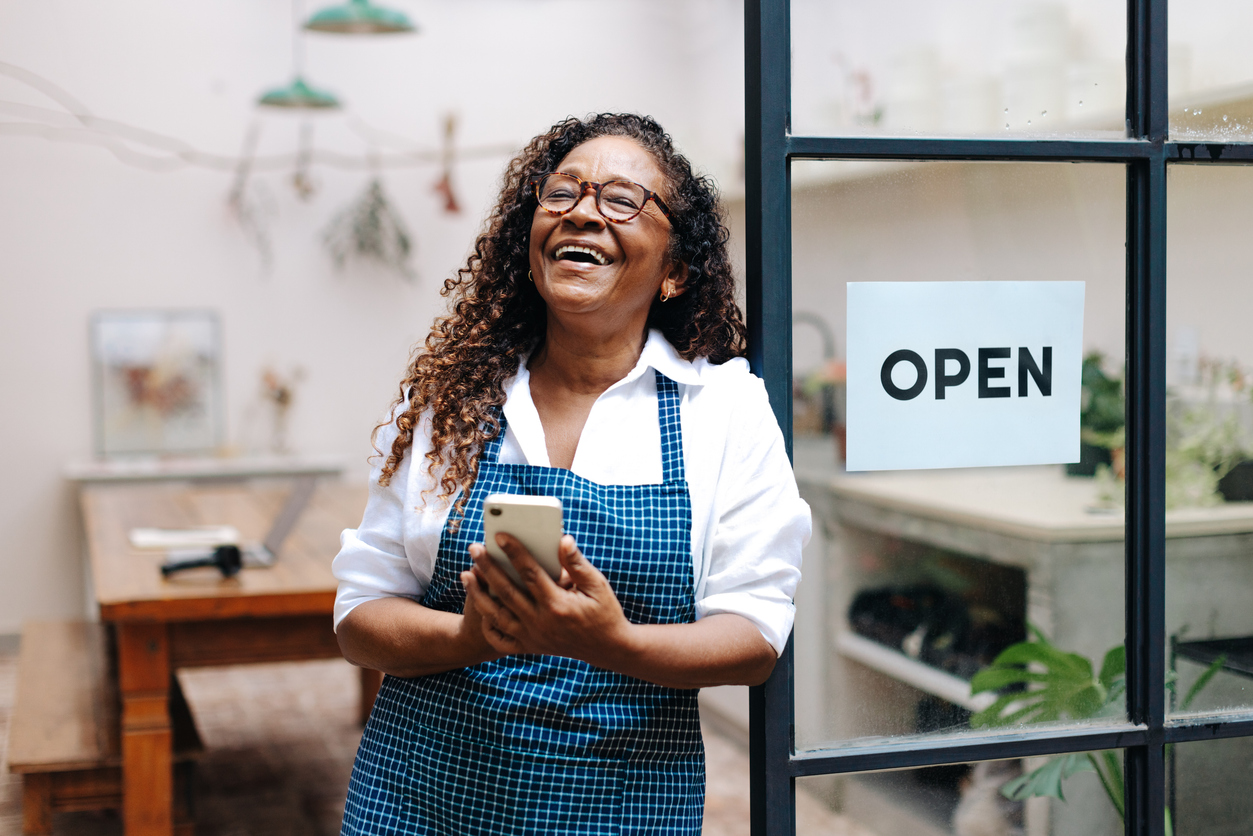 A business owner stands in the doorway of her small business, smartphone in hand. She is smiling. The Valley List offers the best cheap web hosting in Westerville Ohio,