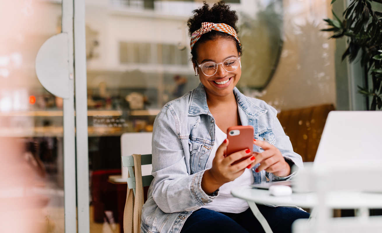 A woman sits in a cafe and smiles as she looks at her phone. The Valley List has partnered with Web Impact Marketing to offer digital marketing solutions.