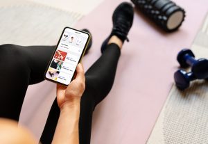 A person in yoga pants and tennis shoes sits on a yoga mat with stretching equipment around them. Their face is not visible, however they are holding their smart phone with a mobile app. The Valley List has partnered with Web Impact Marketing to offer digital marketing solutions.