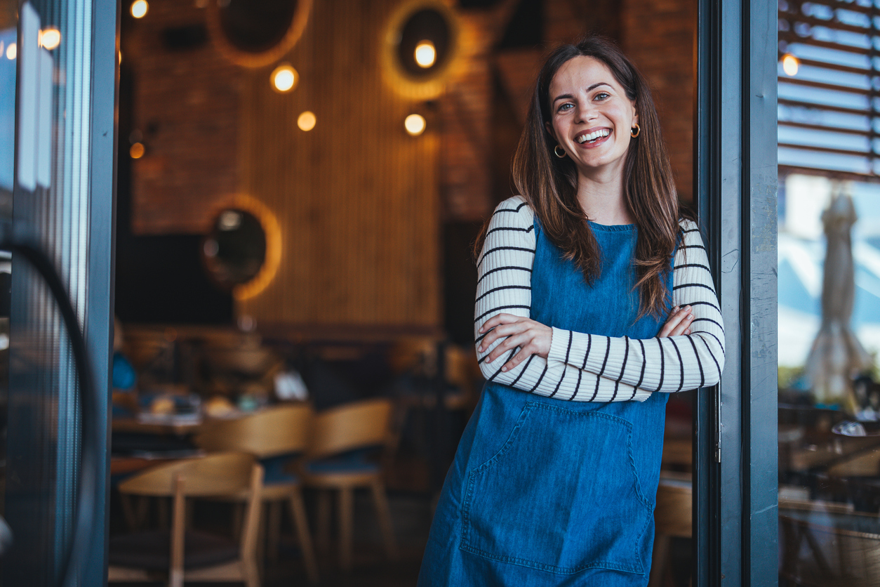 A woman in a denim jumper smiles at the door of her coffee shop. She has a striped shirt on and long brown hair. The Valley List provides affordable SEO in Cambridge Ohio.