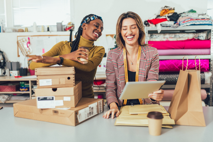 two women work in their small business, packing orders together. One woman looks over the other person's shoulder at a tablet. The Valley List is a website design company in Worthington Ohio who will meet all of your design and development needs.