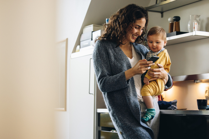 A smiling woman stands in her kitchen with her baby in one arm, and her cell phone in the other, she and the baby both smile and look at screen. The Valley List offers app development in Worthington, Ohio.