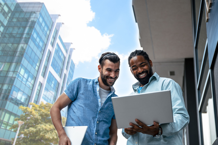 Two young men stand outside with a tall building in the background. They are looking at a laptop together. The Valley List and Web Impact Marketing have partnered to become the best digital marketing agency in Dublin, Ohio.