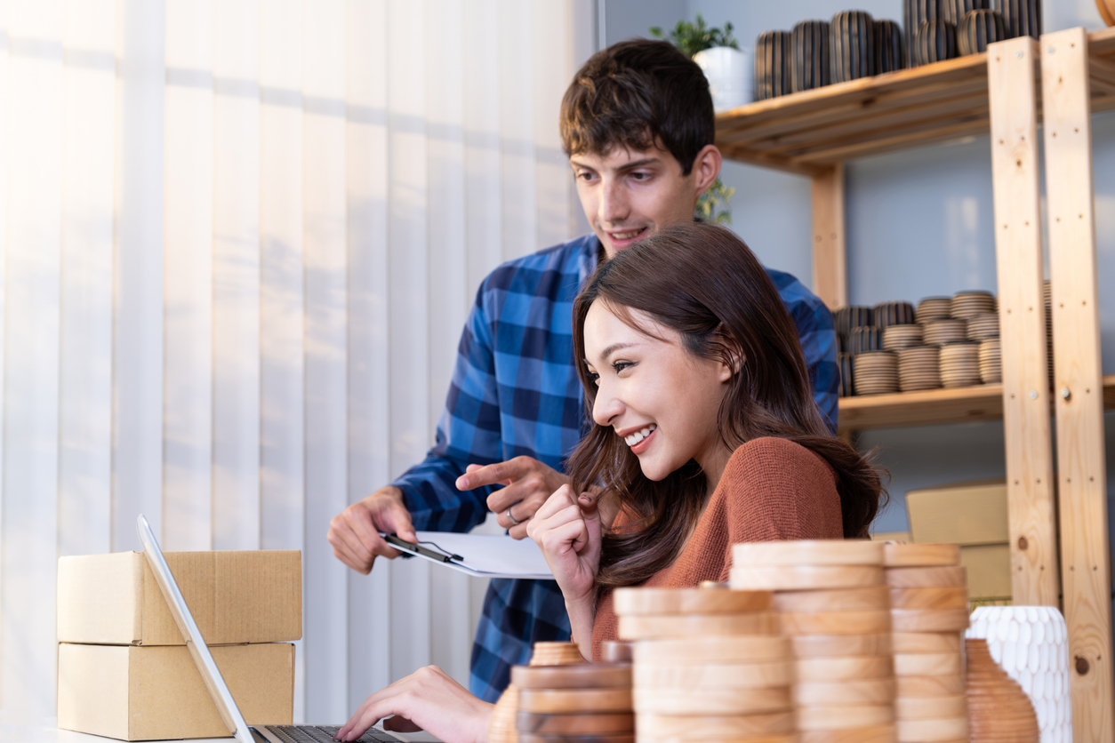A young business owner sits at her laptop, smiling. She is surrounded by her wooden coasters and other products, while a man looks over her shoulder at the screen, helping her. The Valley List is here to help build affordable and mobile website design in Westerville, Ohio.