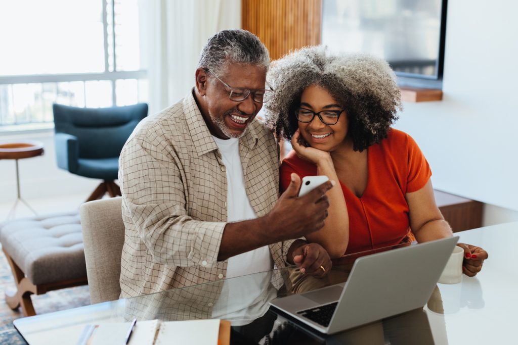 A smiling couple uses technology, including a laptop and cellphone in their home. Chrome accessibility settings help everyone, from every walk of life.