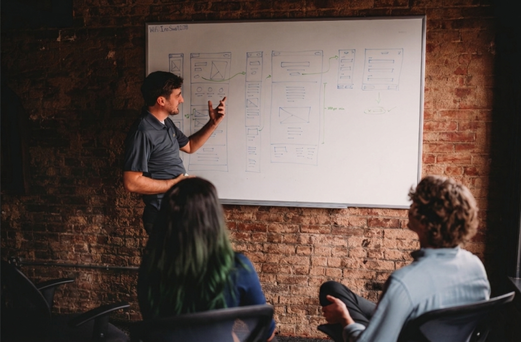 A man points to a whiteboard with a wireframe, while two people watch and learn.